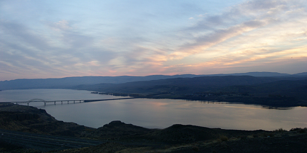 [Two images stitched together to show the broad expanse of land as viewed from above the bridge over the Columbia River as the sun sets creating pink streaks in the sky.]